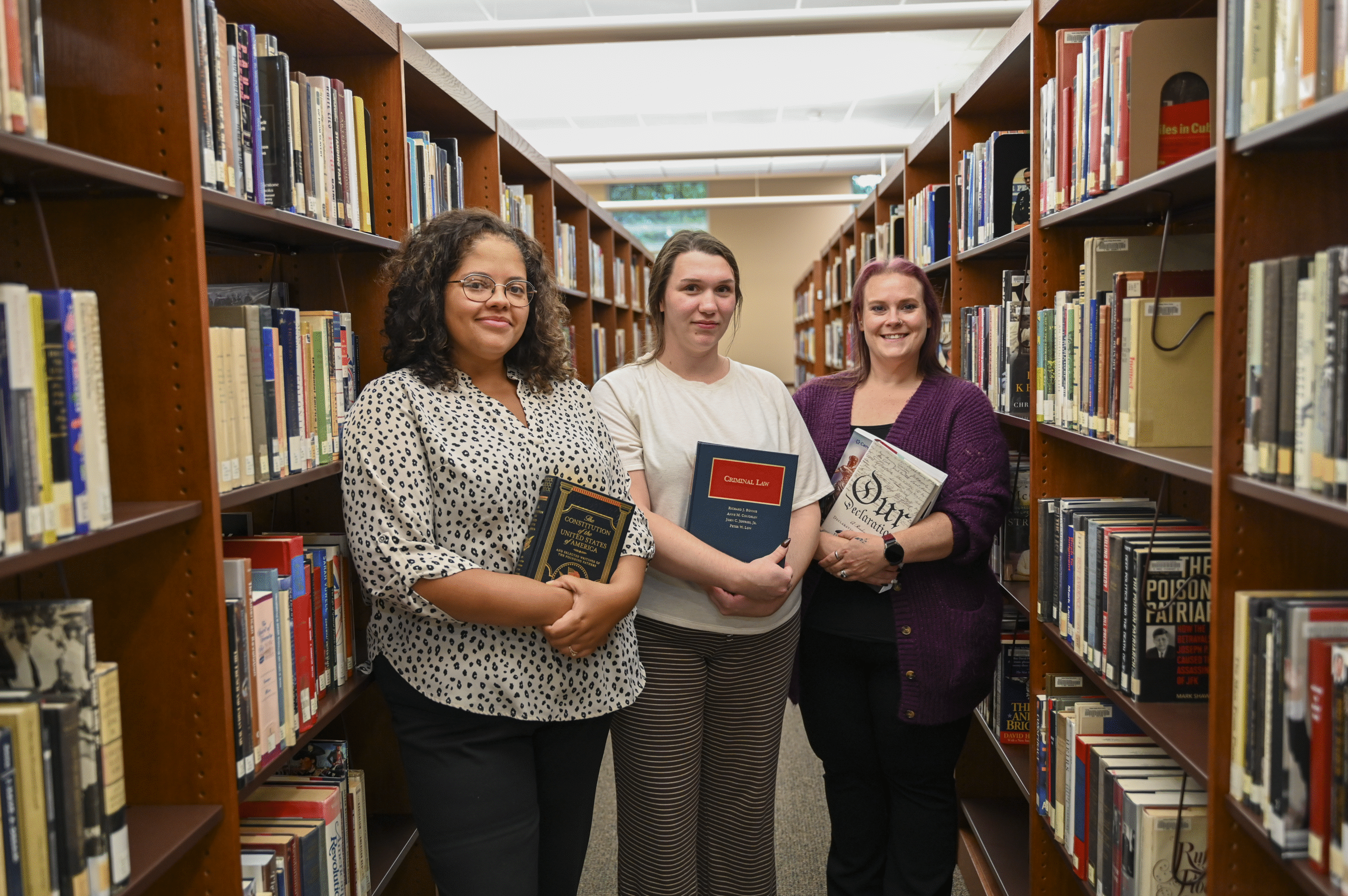 Three female alumni standing in the library holding law books.